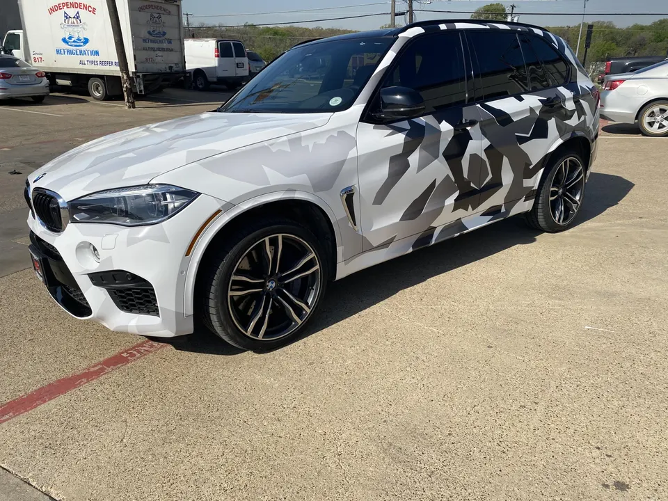 A white BMW X5 SUV with a black and white camouflage wrap parked outdoors, showcasing its custom exterior and black wheels.