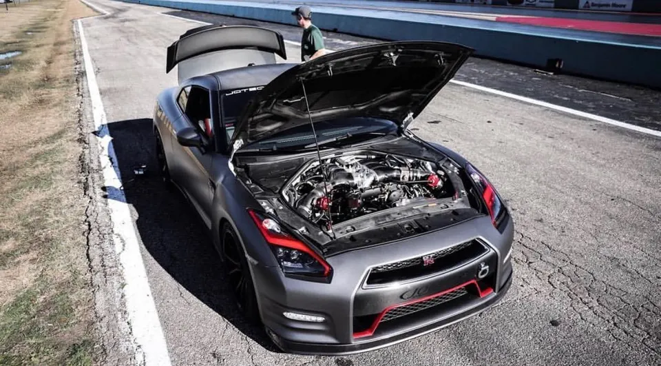 Matte grey Nissan GT-R with its hood and trunk open, showcasing the engine bay on a drag strip with a person standing by.