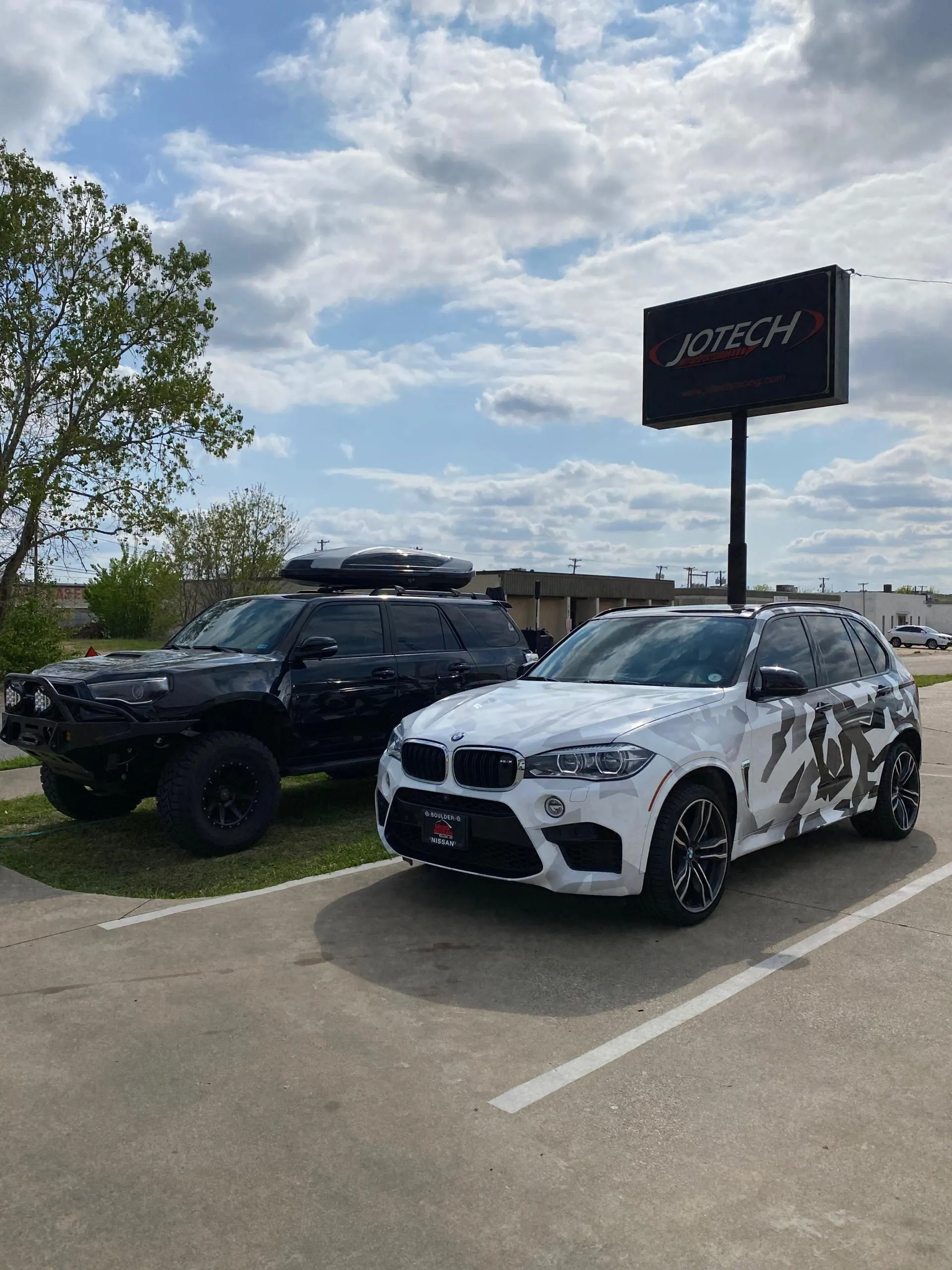 Two modified SUVs, a black off-road vehicle and a white BMW X5 M with a camouflage wrap, parked outside Jotech Motorsports.