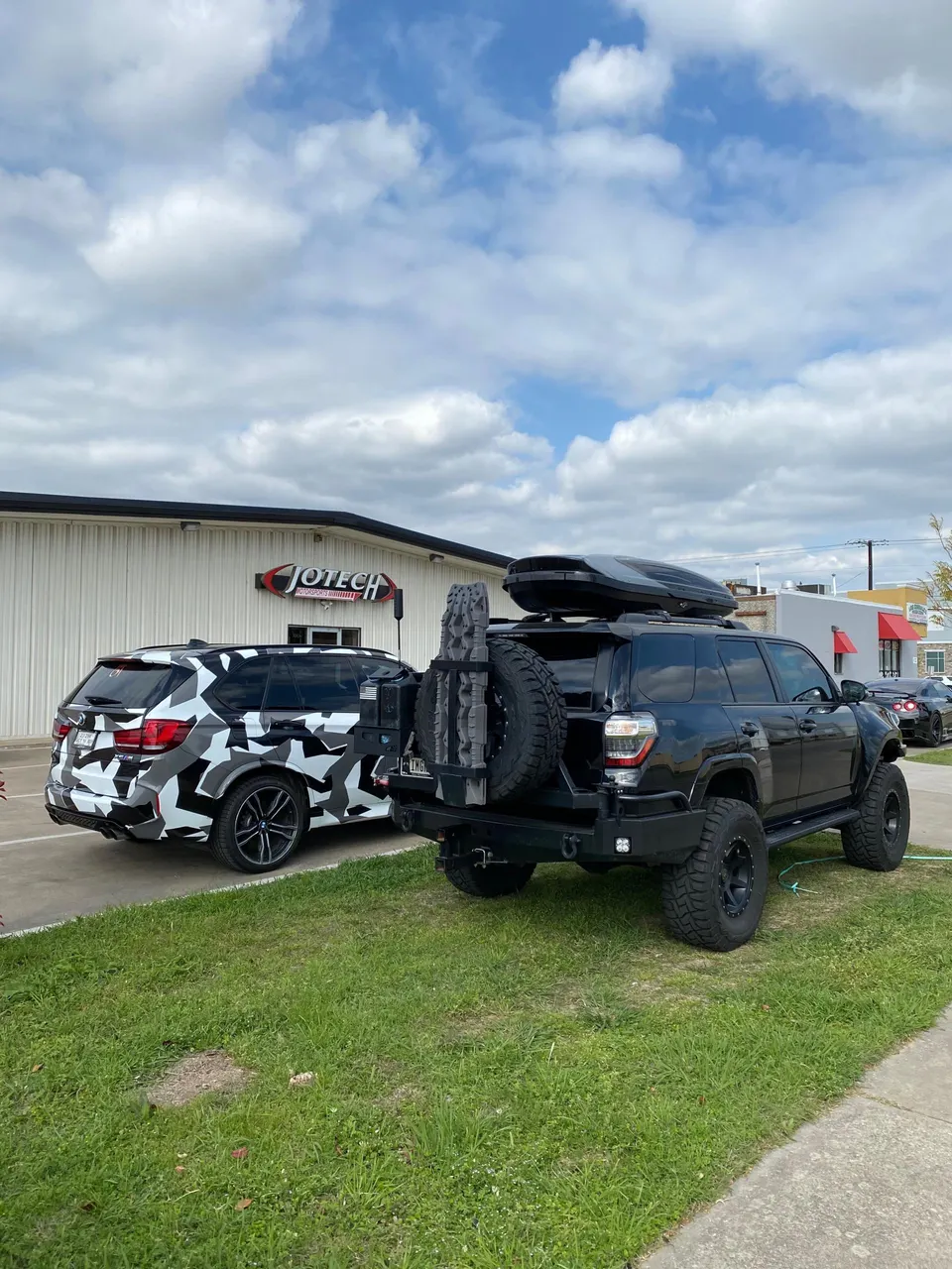 A black Toyota 4Runner with off-road modifications and a BMW X5 with a camouflage wrap parked outside Jotech Motorsports.