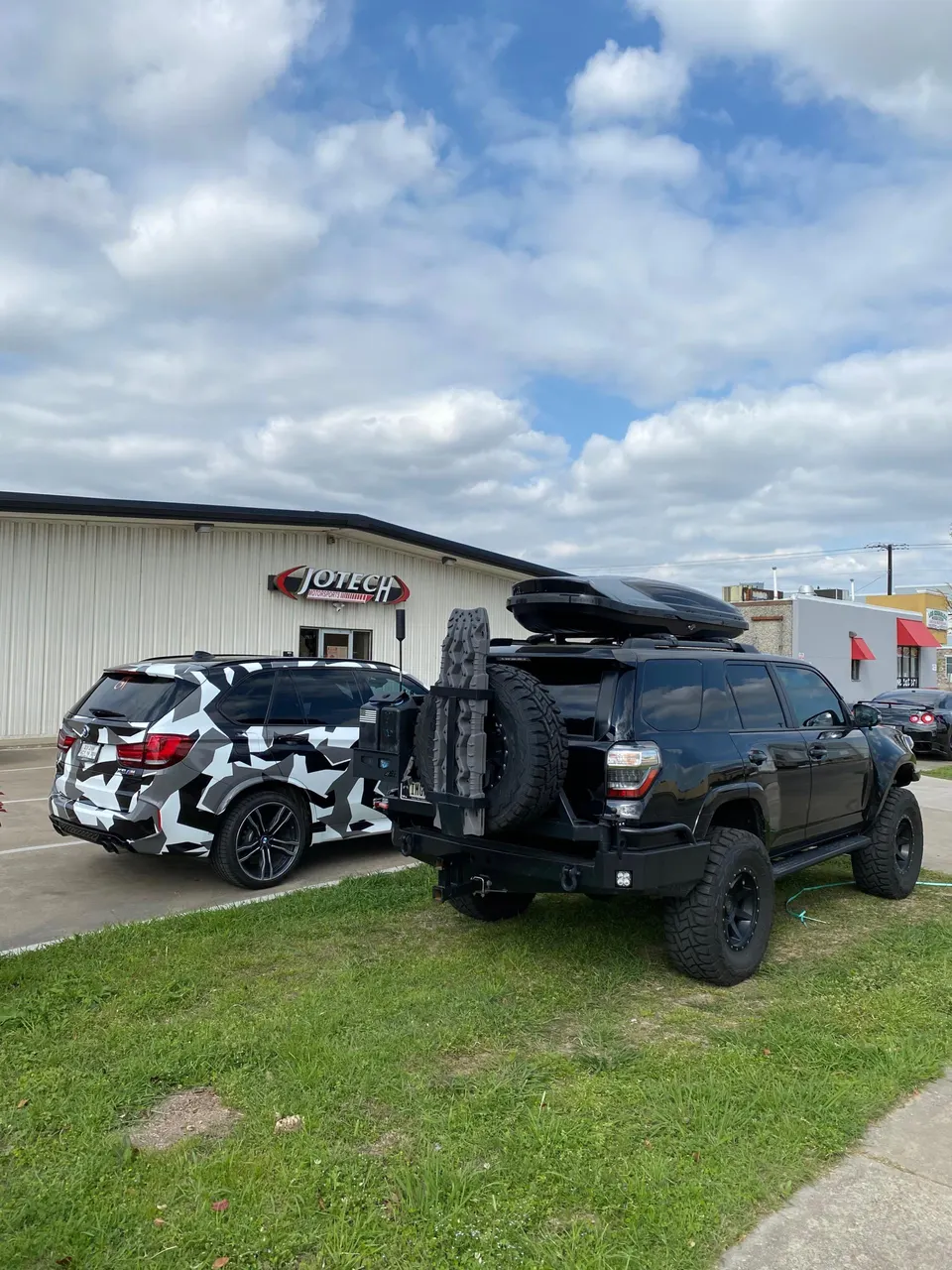 A black Toyota 4Runner with off-road modifications and a white BMW X5 with a camouflage wrap parked outside Jotech Motorsports.