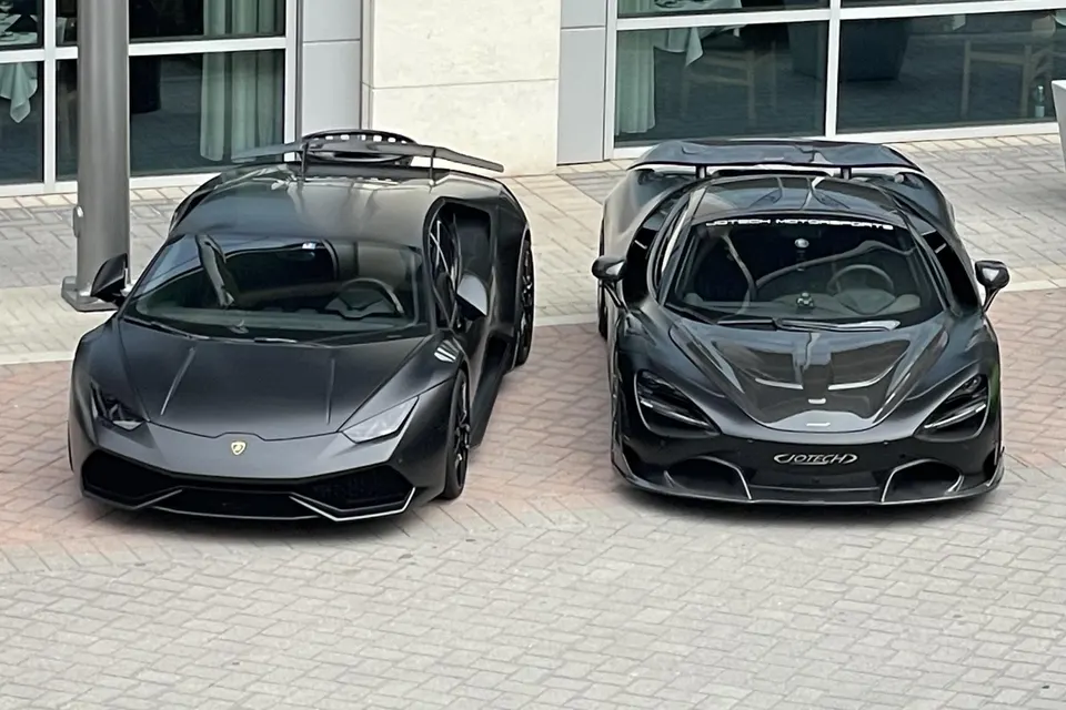 Two matte black supercars, a Lamborghini Huracan and a McLaren 720S, parked side-by-side outside a building.