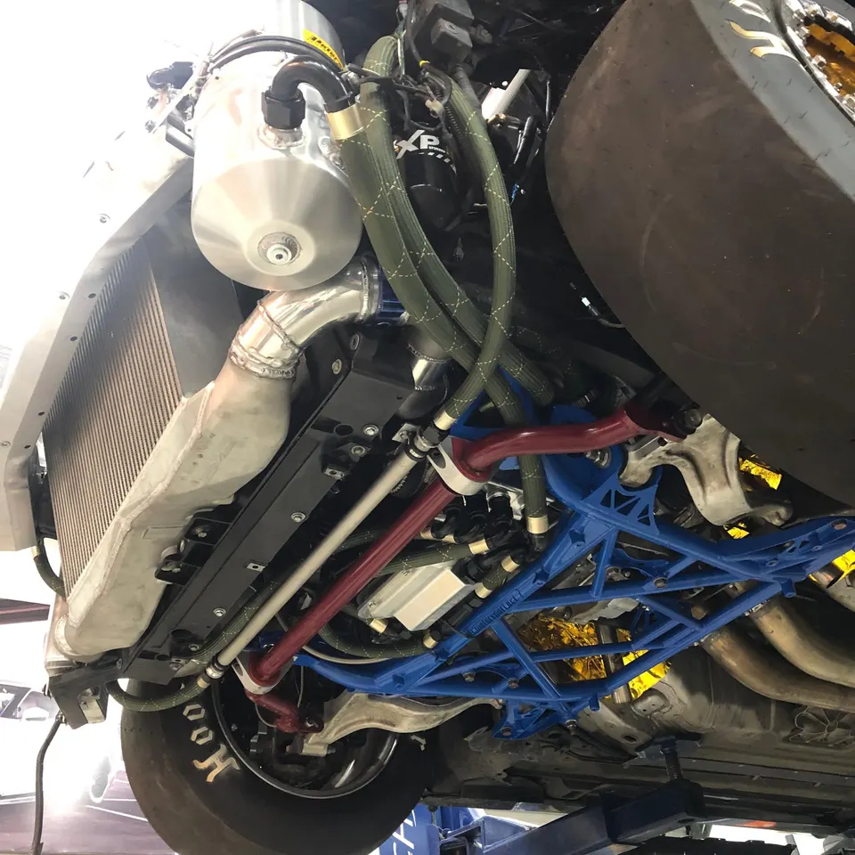 Underside view of a high-performance race car on a lift, showcasing a large intercooler, custom boost piping, and blue subframe.
