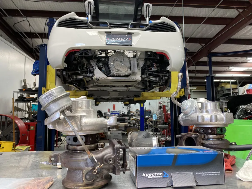 A white McLaren sports car on a lift in a performance tuning shop, with turbochargers and Injector Dynamics boxes on a workbench.