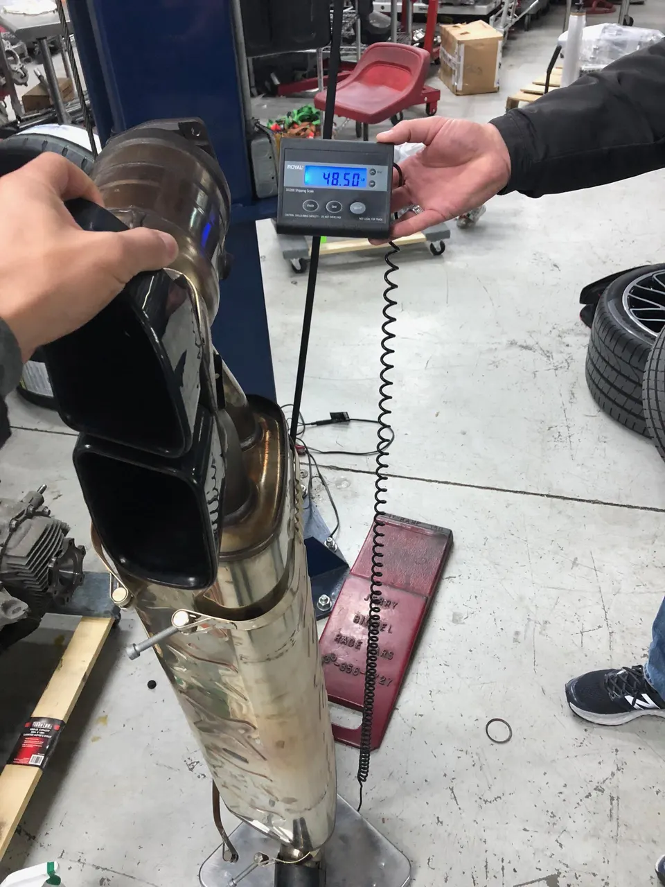 A person weighing a large aftermarket exhaust muffler with dual tips on a digital scale in a performance shop.