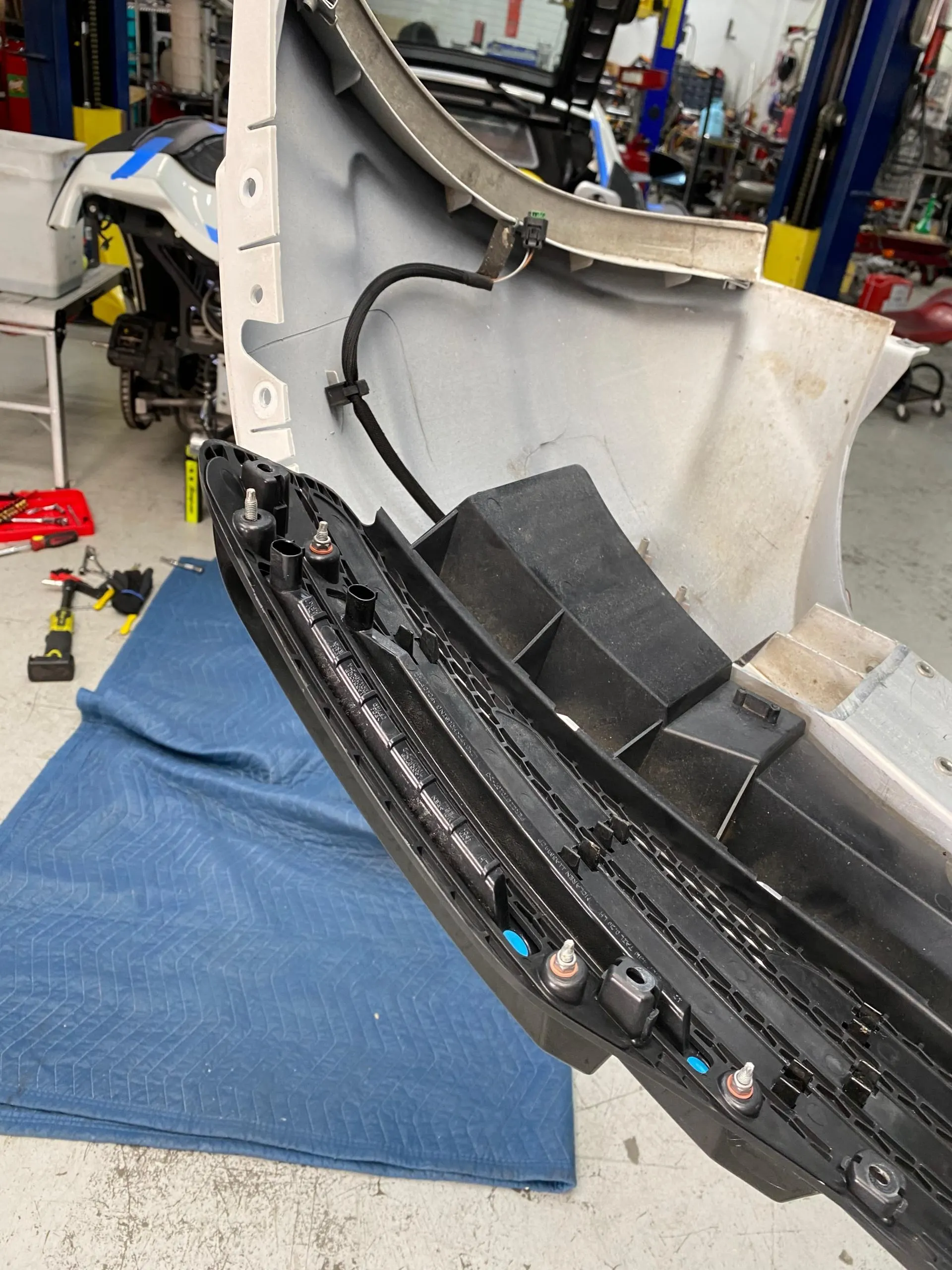 A car bumper removed for service in a professional auto shop, showing internal mounting points and wiring.