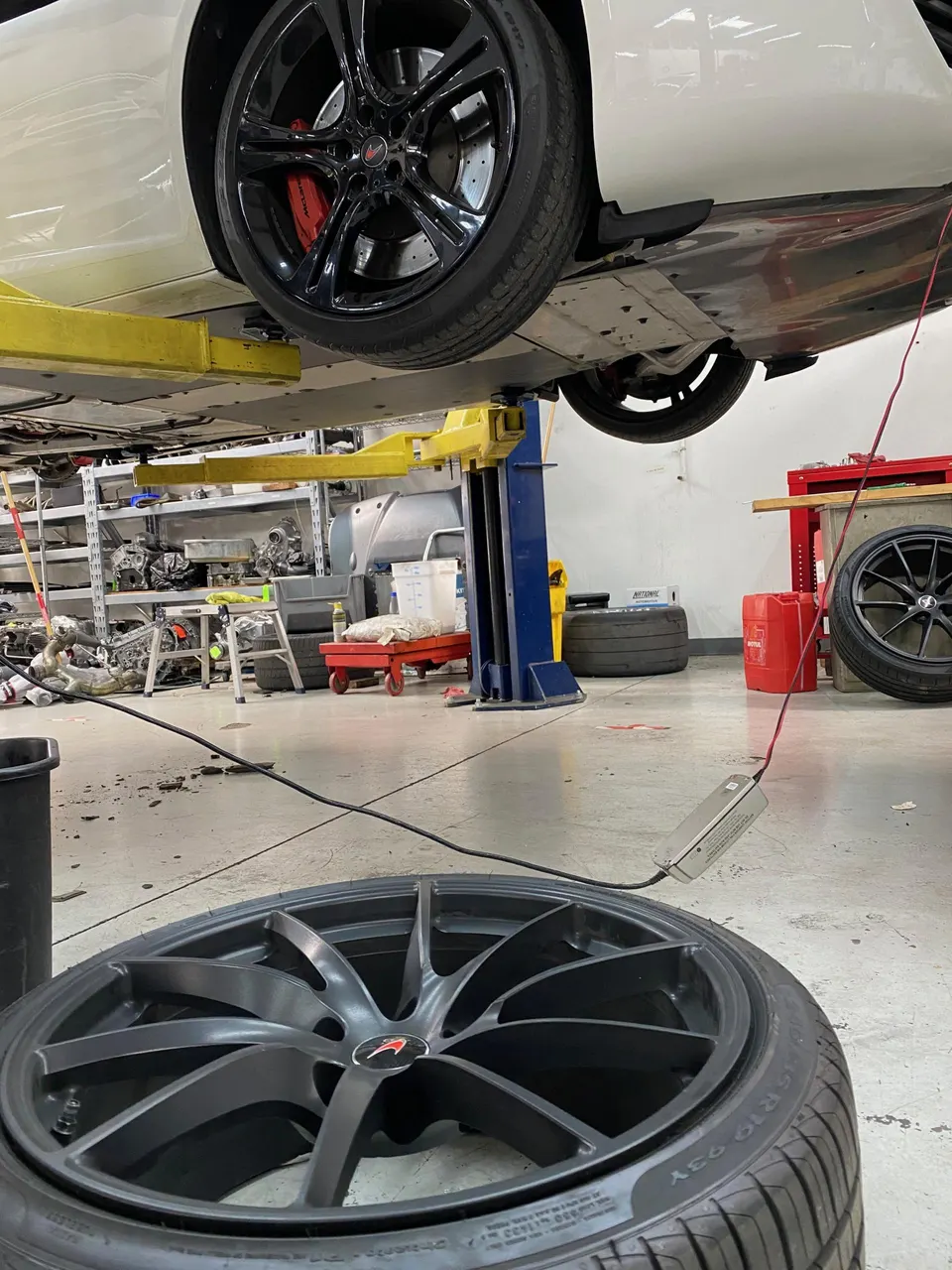 A white McLaren supercar on a lift in a performance tuning shop, with a wheel and red brake calipers visible.