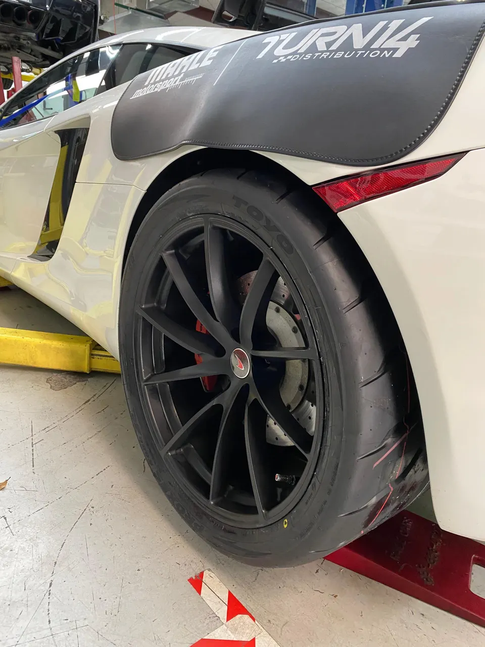 A white McLaren on stands in a shop, showcasing its black aftermarket wheels, Toyo tires, and red brake calipers.