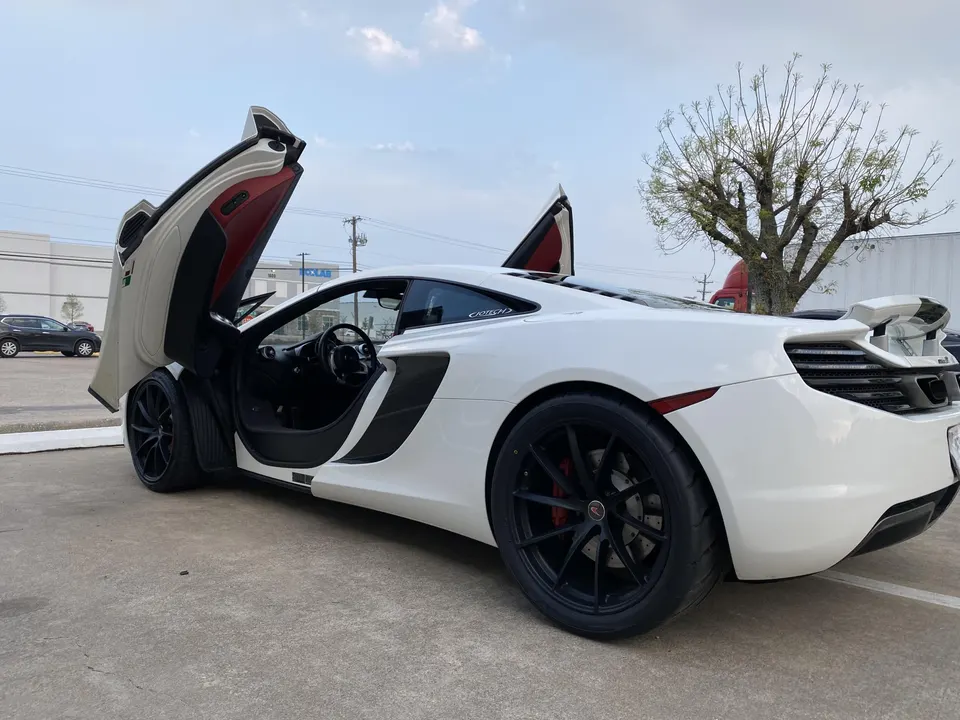 White McLaren supercar with its distinctive doors open, parked outdoors on a sunny day.