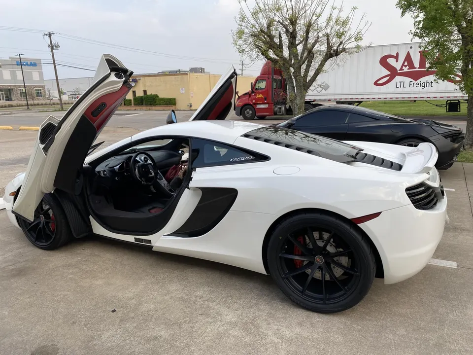 White McLaren with butterfly doors open parked next to a black McLaren in an outdoor lot.