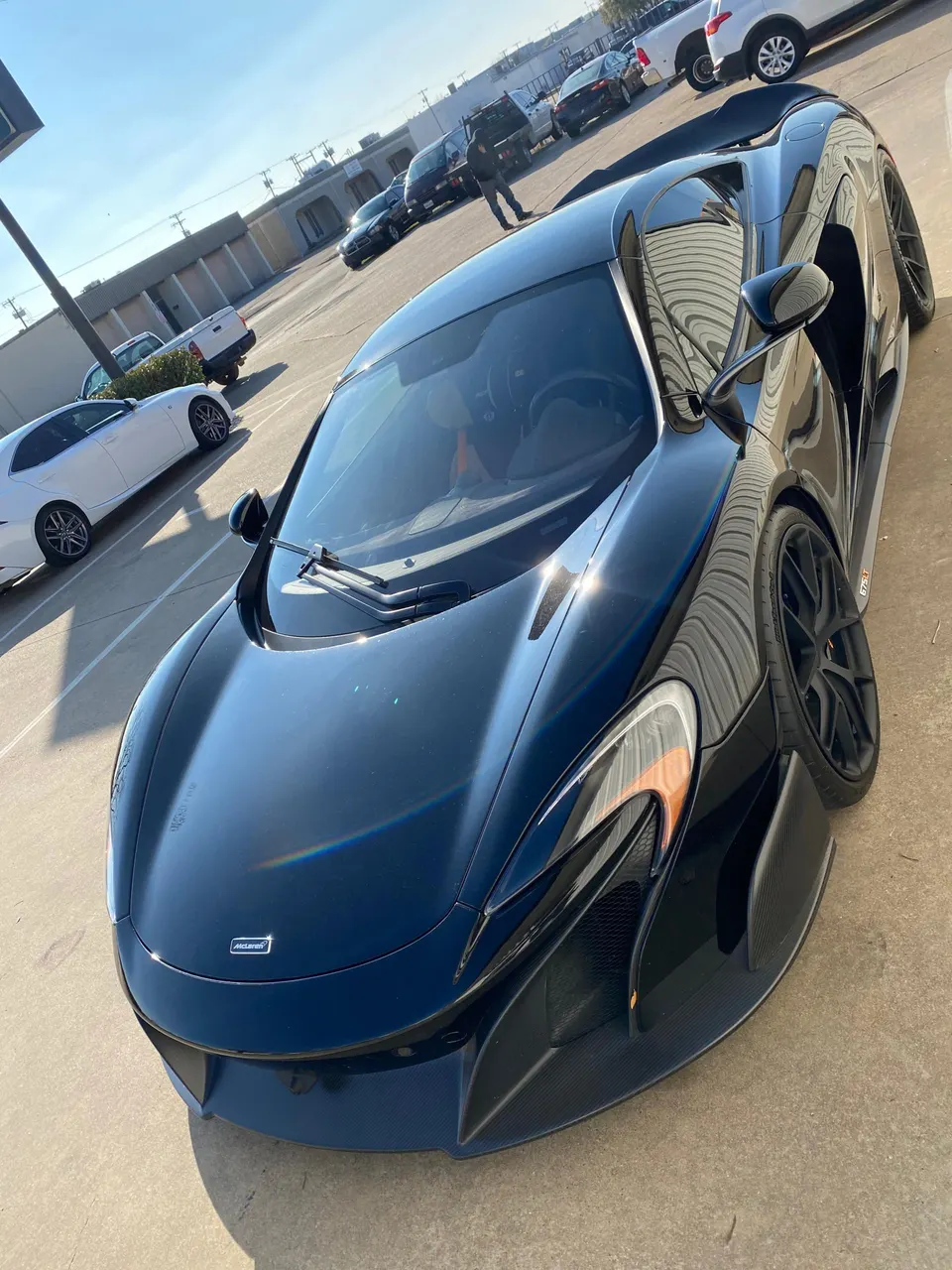 A black McLaren supercar parked in an outdoor lot on a sunny day, showcasing its sleek design and carbon fiber accents.