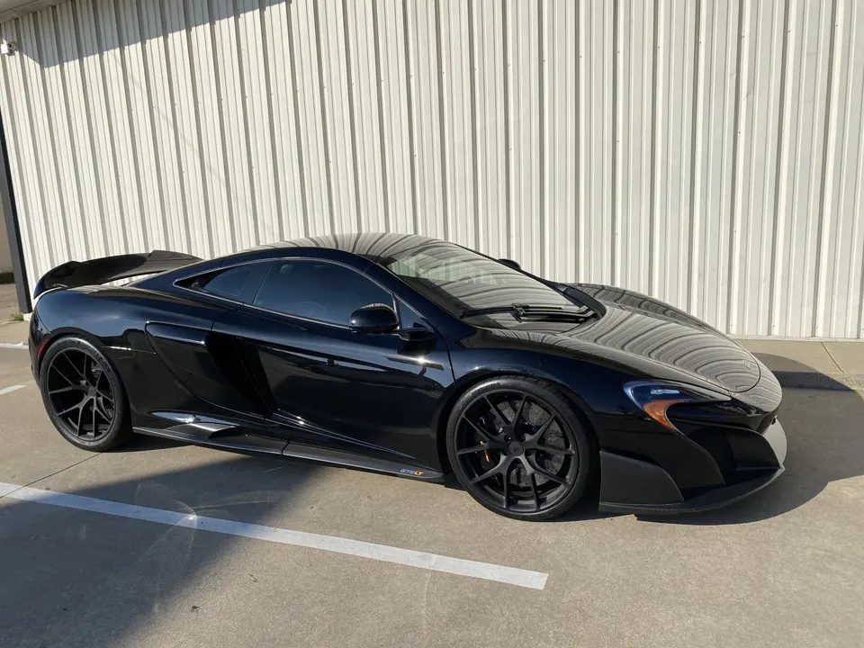A black McLaren 675LT sports car with aftermarket wheels and carbon fiber aero parked outdoors next to a white wall.