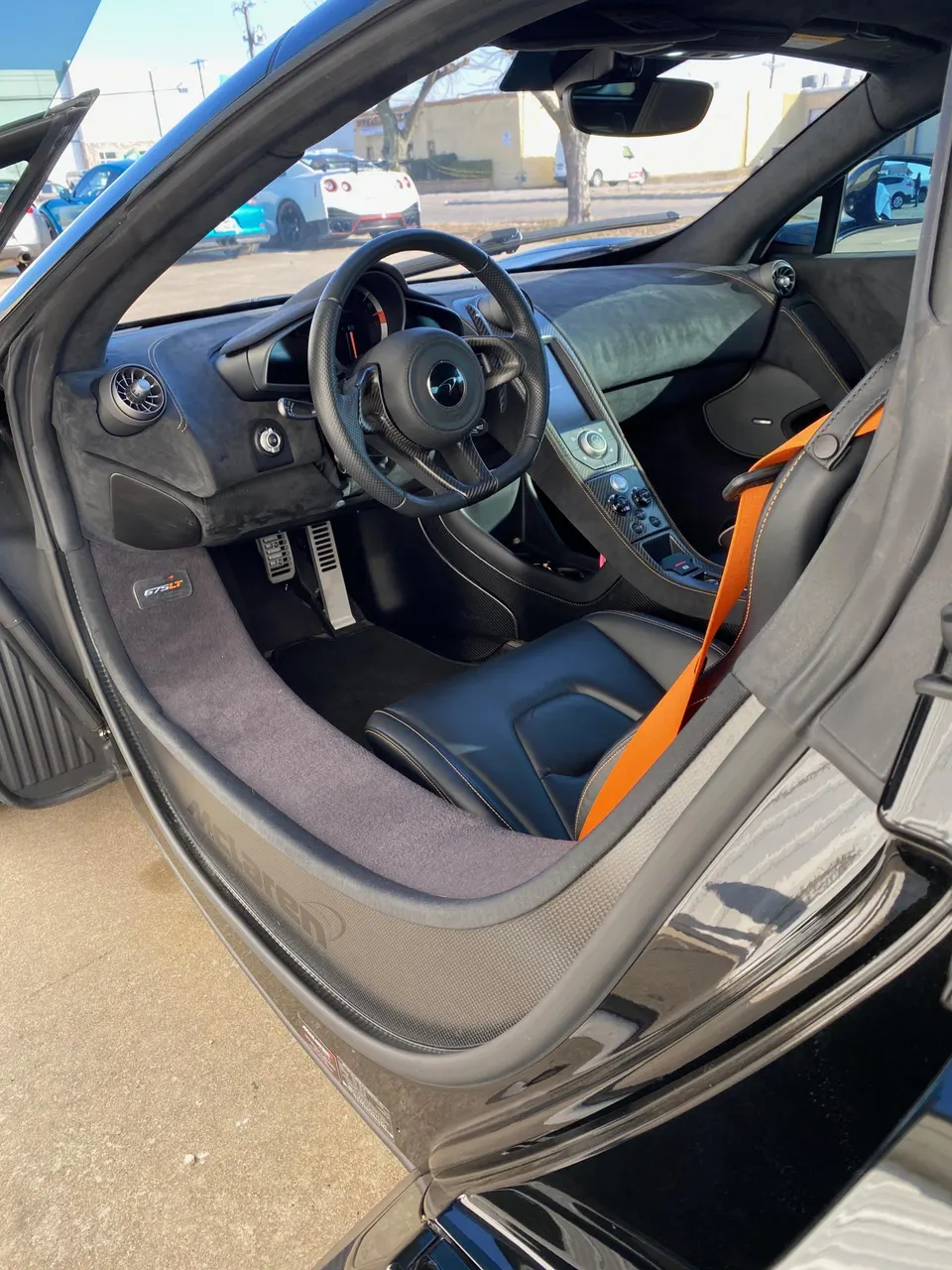 Interior view of a black McLaren 675LT showing the dashboard, carbon fiber steering wheel, and orange seatbelt.
