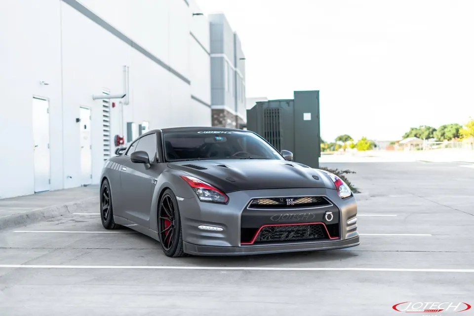 A grey modified Nissan GT-R R35 with a black carbon hood and red accents parked in a lot outside a modern building.