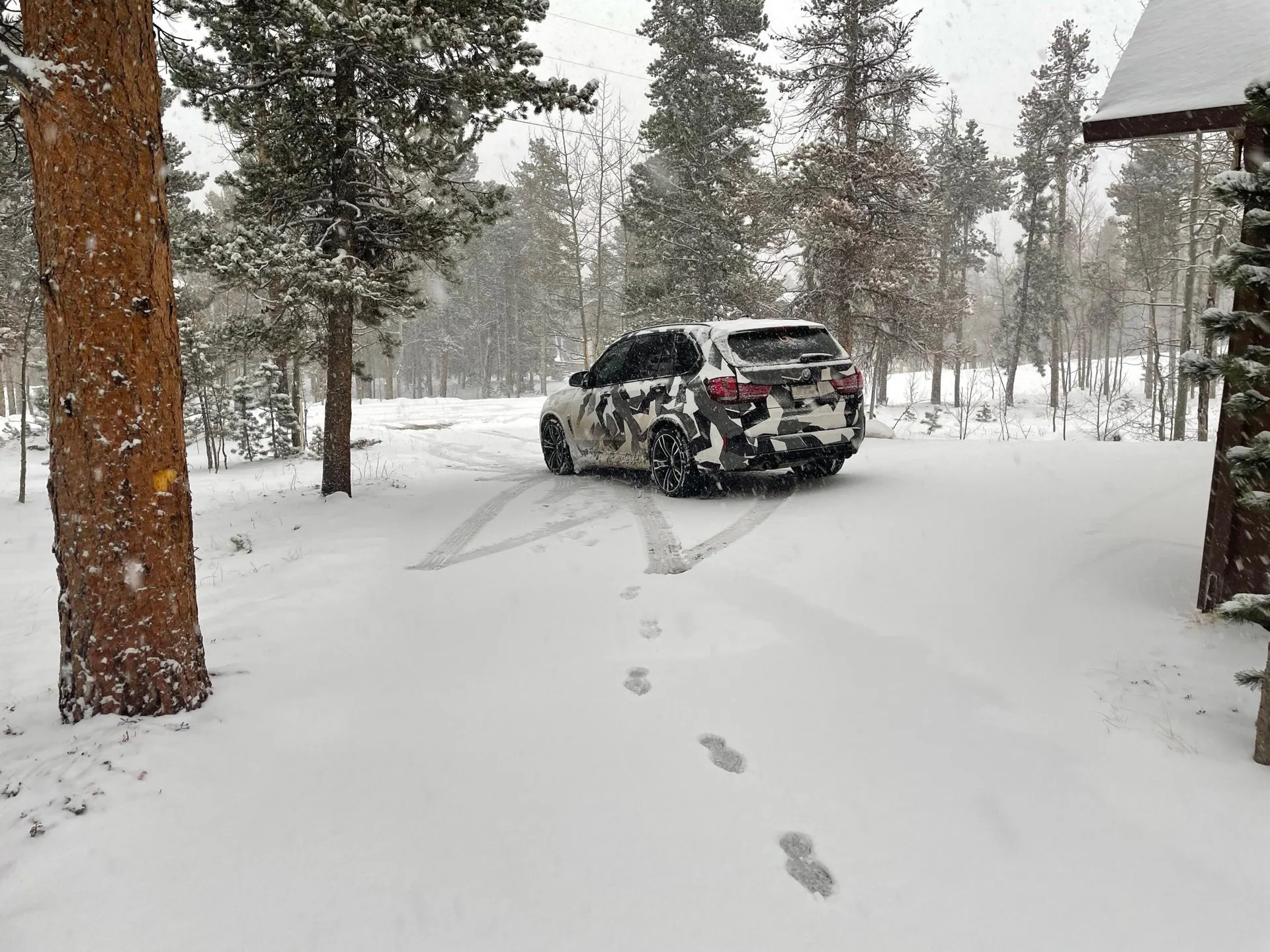A BMW X5 SUV with a camouflage wrap parked in a snowy forest, with tire tracks and footprints in the snow.