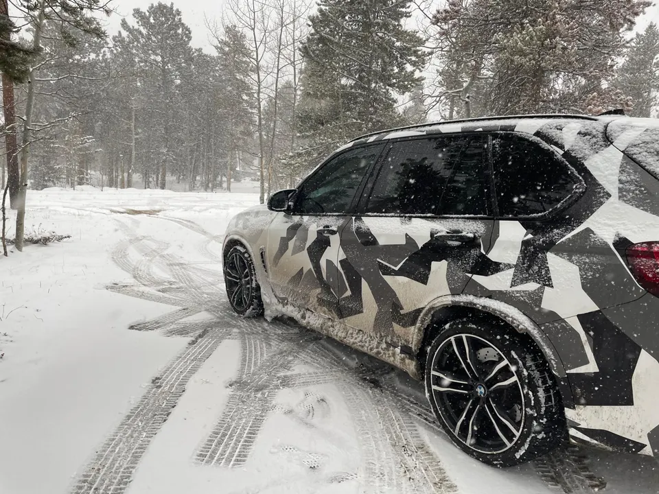 A BMW X5 SUV with a distinctive camouflage wrap parked in a snowy forest, tire tracks visible in the fresh snow.