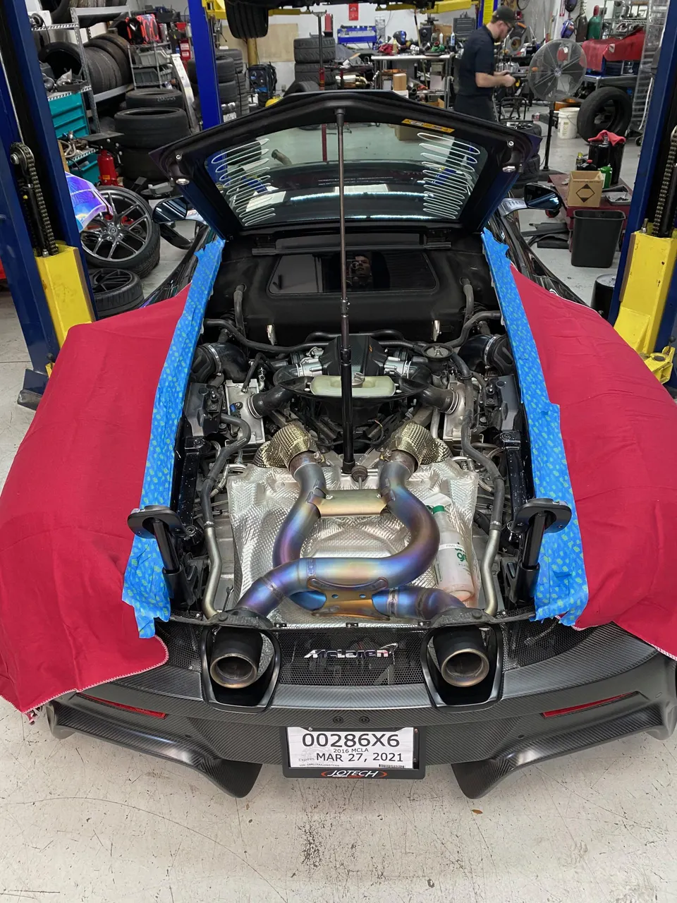 A McLaren supercar with its engine bay open, showcasing a vibrant titanium exhaust system and air filters in a performance tuning shop.