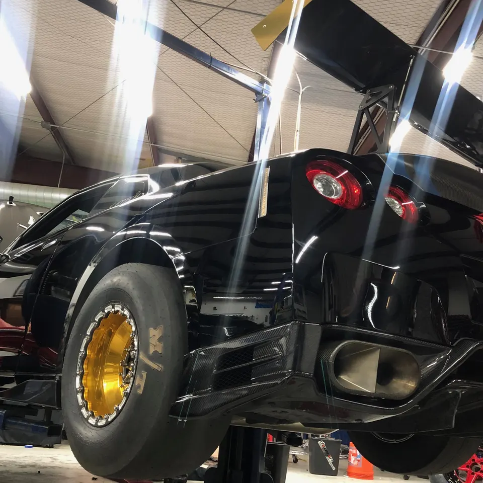 A black Nissan GT-R drag car on a lift in a shop, featuring a large carbon fiber rear wing, diffuser, and gold drag wheels.