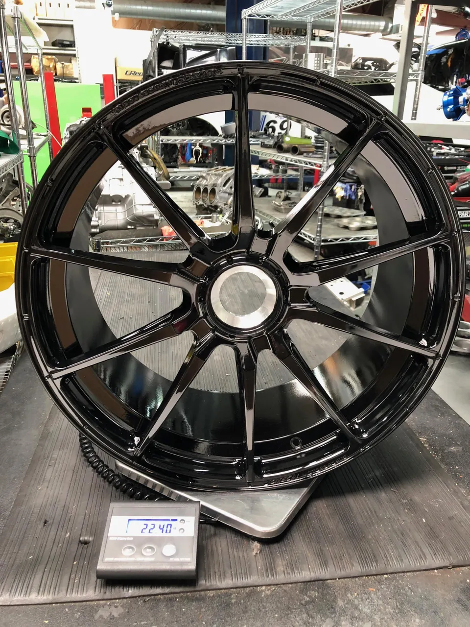 Black multi-spoke performance wheel being weighed on a digital scale in a shop environment.