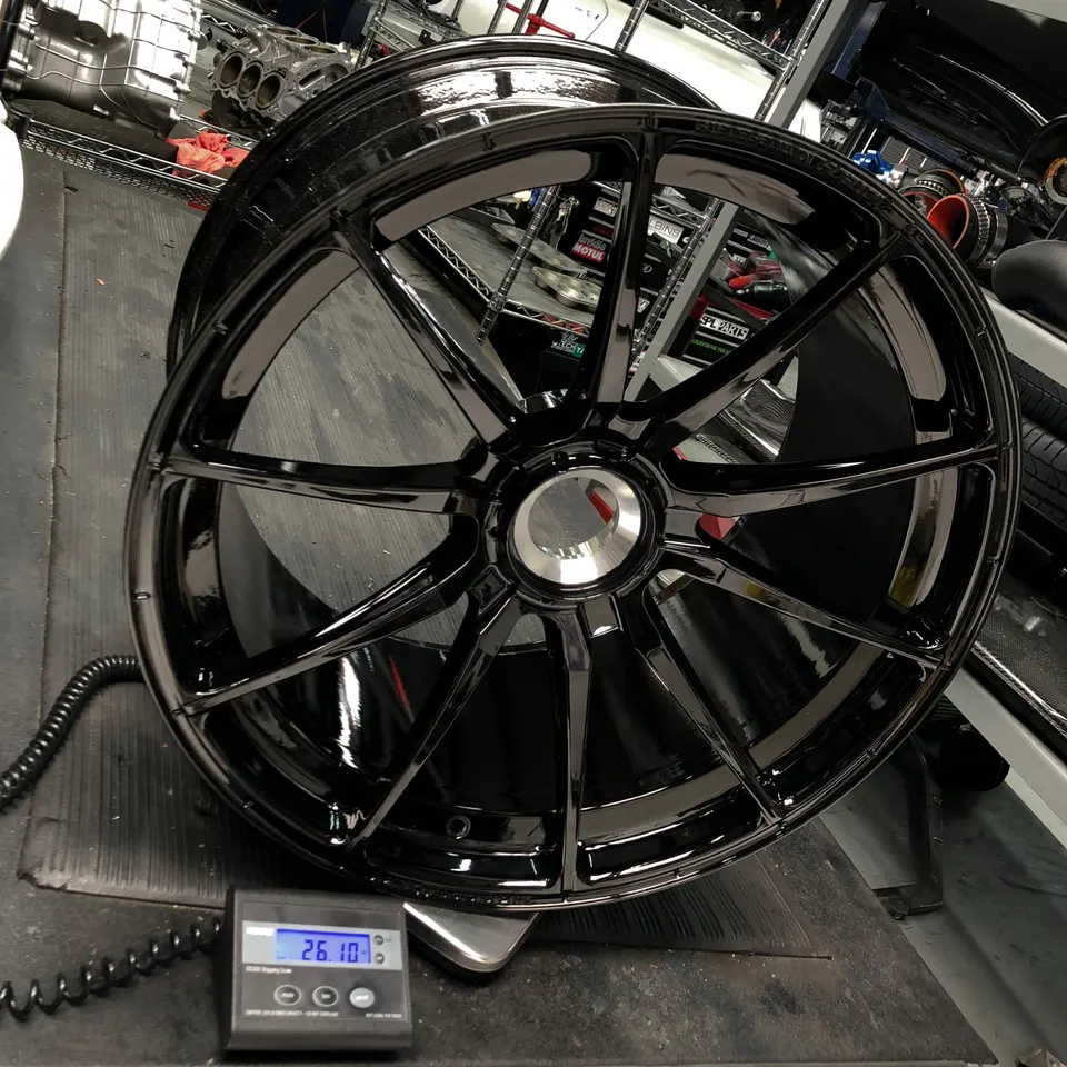A glossy black multi-spoke performance wheel being weighed on a digital scale in a Jotech Motorsports shop.