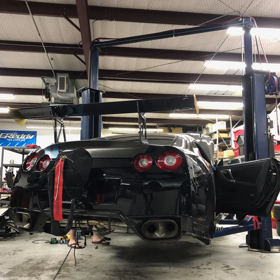A black Nissan GT-R on a lift in a performance shop, featuring a large carbon fiber rear wing and aftermarket exhaust.