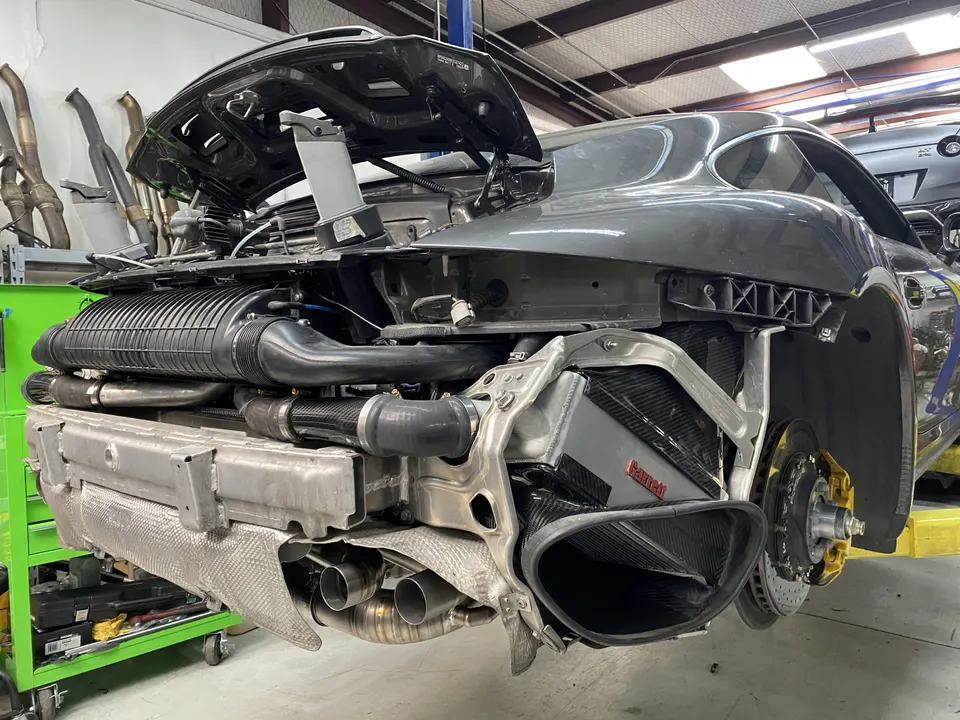 A Porsche 911 on a lift in a shop with its rear bumper removed, exposing the intercooler, exhaust, and engine components.