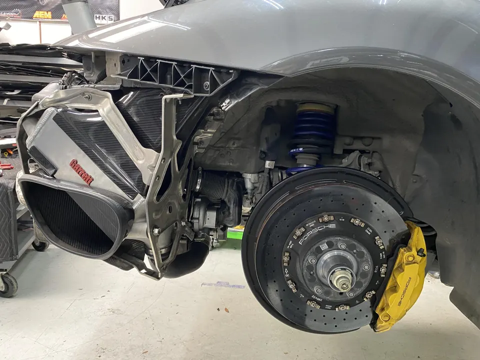 A Porsche car in a shop with its front fender removed, showcasing a carbon fiber Garrett intake, blue coilover suspension, and yellow Porsche brakes.