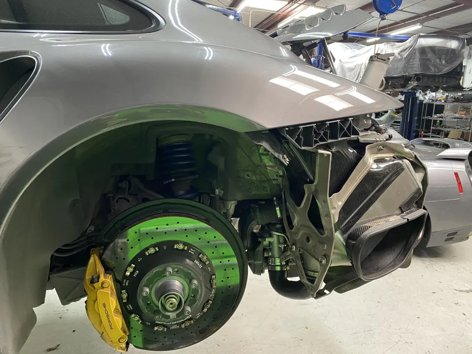 A grey Porsche 911 in a shop with the rear bumper removed, showcasing yellow brake calipers, drilled rotors, and suspension.