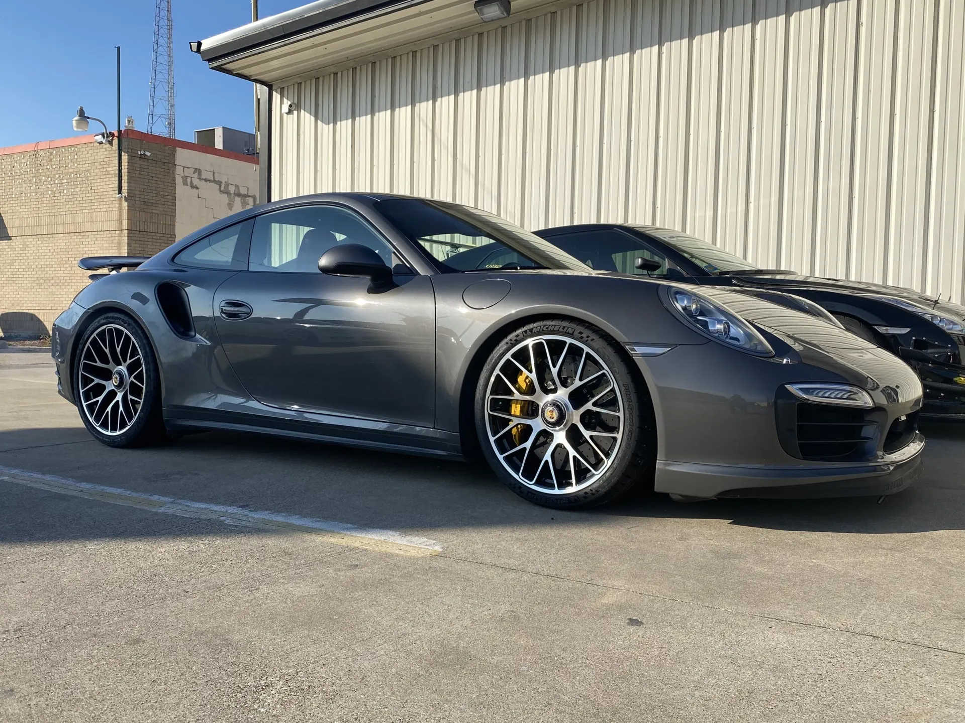 A grey Porsche 911 Turbo S with aftermarket wheels and yellow brake calipers parked outside a building.