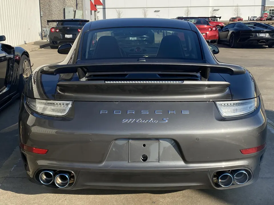 Rear view of a grey Porsche 911 Turbo S with quad exhaust tips and a prominent rear wing, parked outdoors.