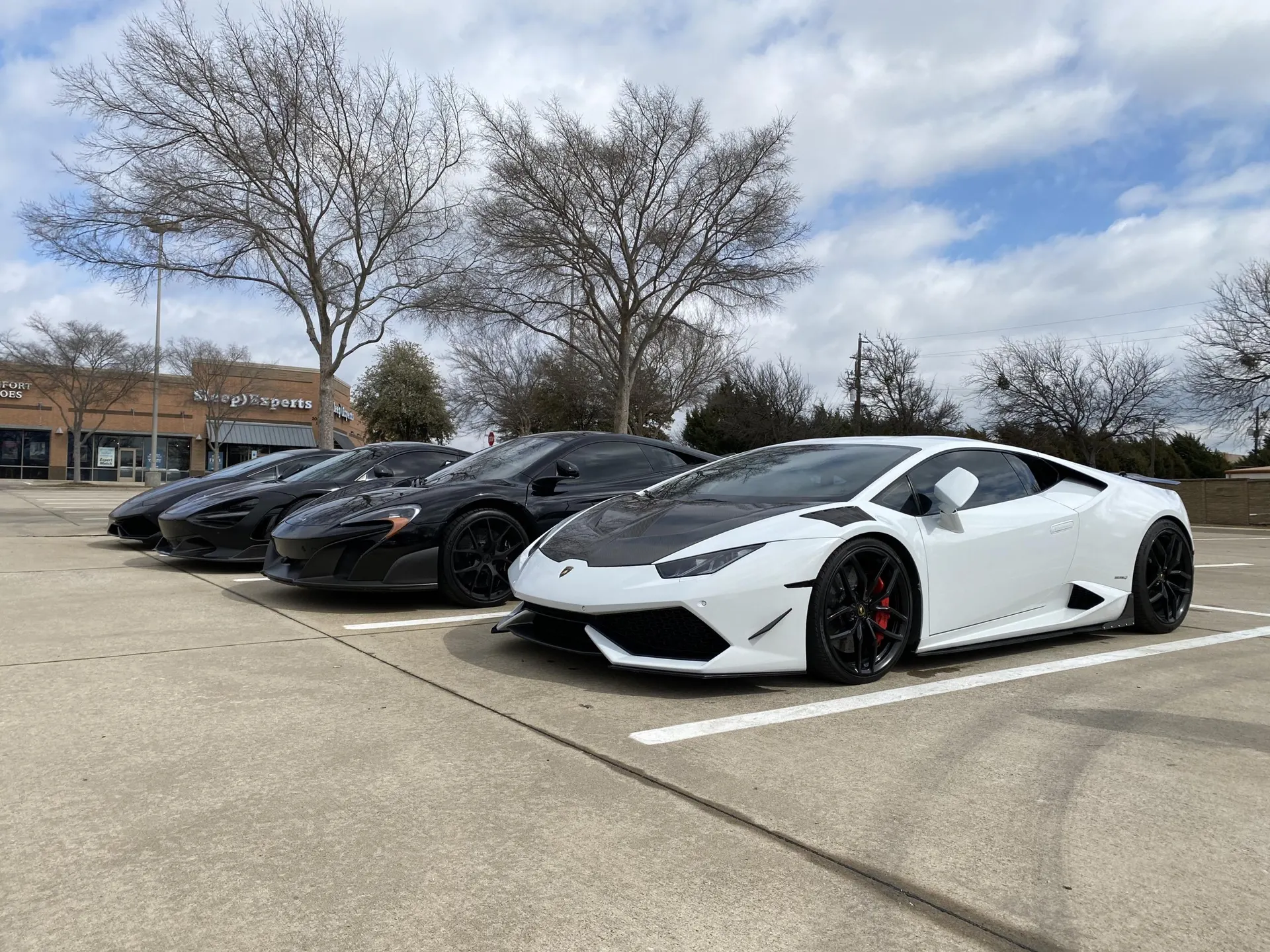A line of supercars, including a white Lamborghini Huracan with a carbon hood and several black McLarens, parked in a lot.