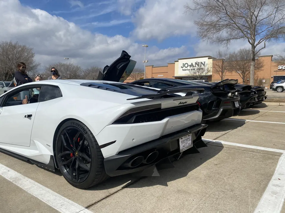 A line of supercars, including a white Lamborghini Huracan and several black McLarens, parked in a shopping center lot with people admiring them.