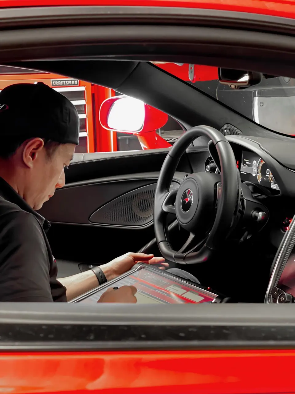 Technician tuning a red McLaren's electronics from the driver's seat in a shop.