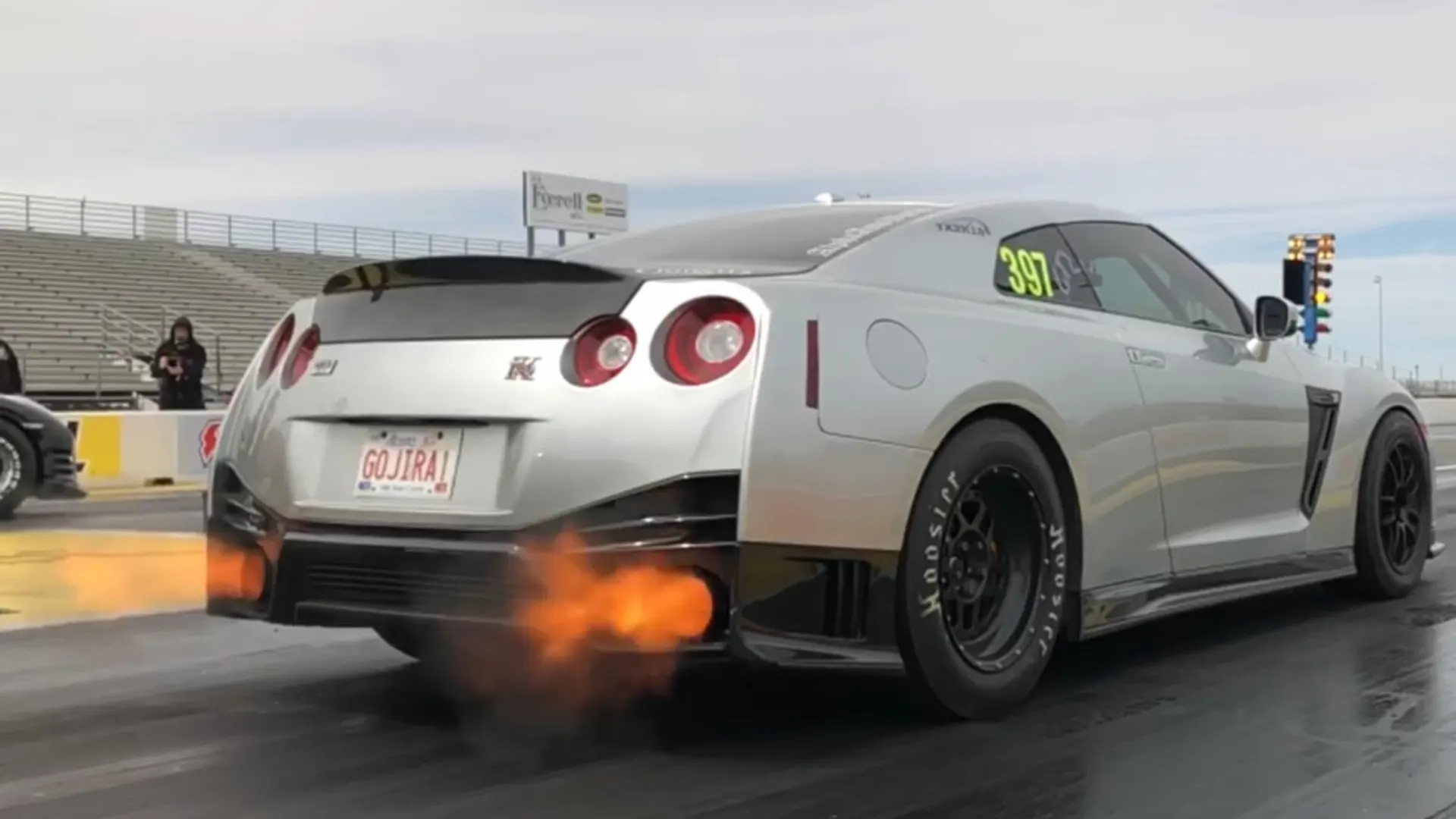 A silver Nissan GT-R launching at a drag strip with flames shooting from the exhaust, showing its rear and side.