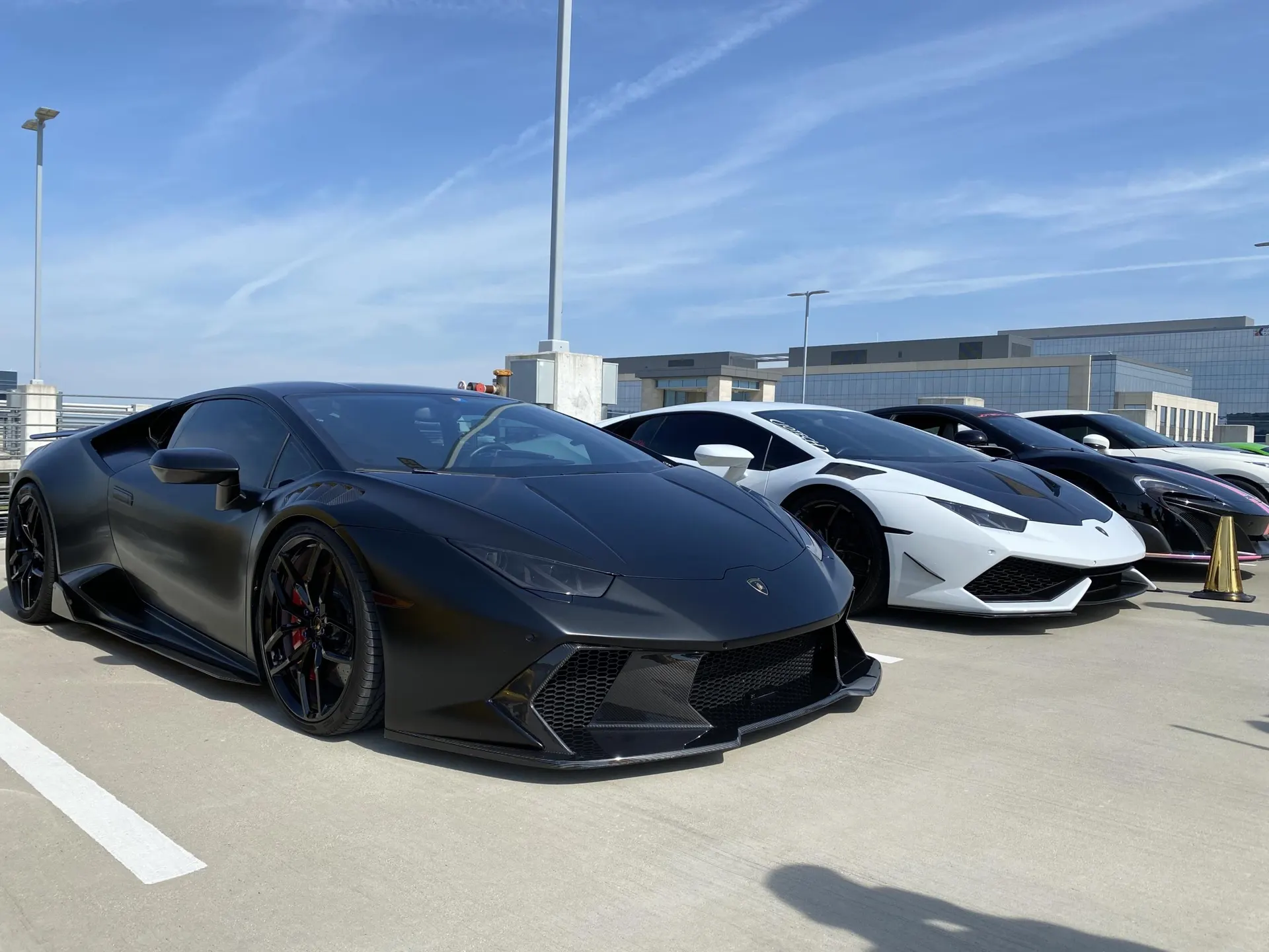 A line of luxury supercars, including a matte black Lamborghini Huracan and a white McLaren, parked outdoors under a clear sky.