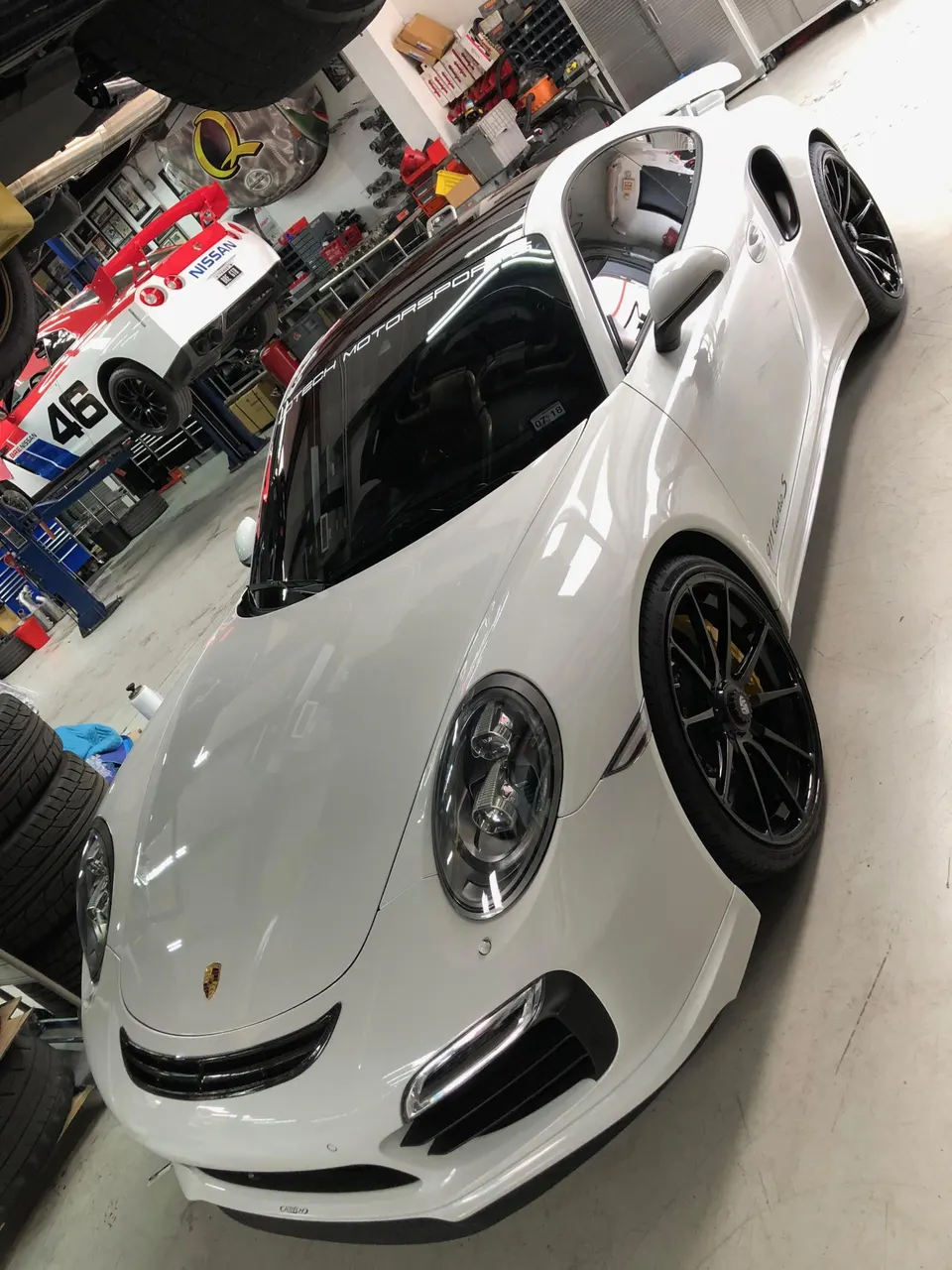 A gleaming white Porsche 911 Turbo S with Jotech Motorsports branding, parked in a workshop with a Nissan GT-R on a lift.