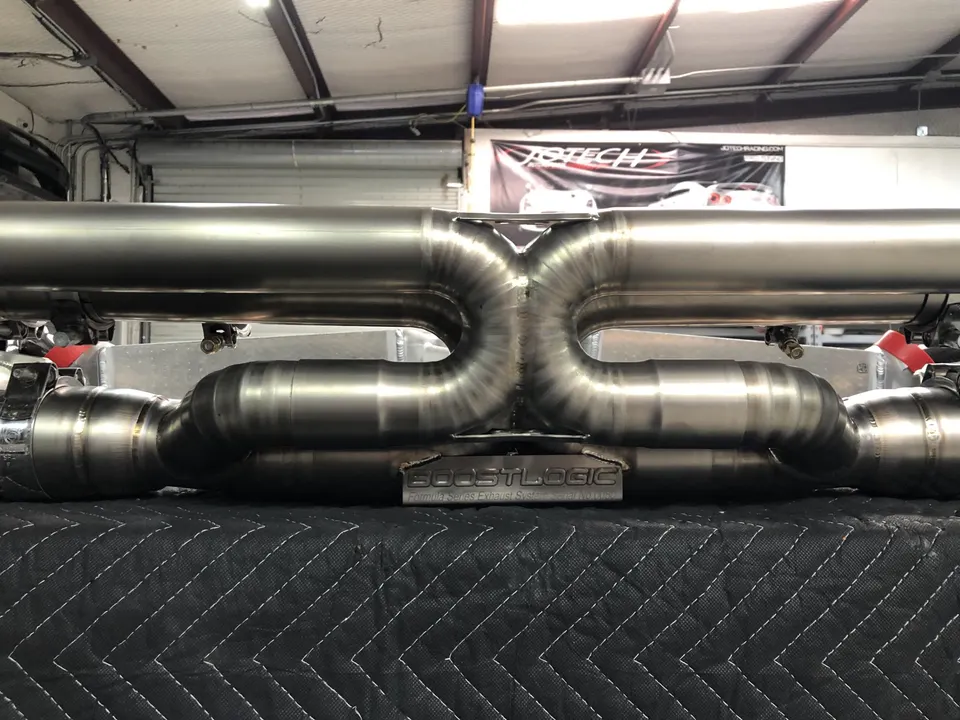 Close-up of a Boost Logic titanium exhaust system with an X-pipe design, displayed in a shop setting.