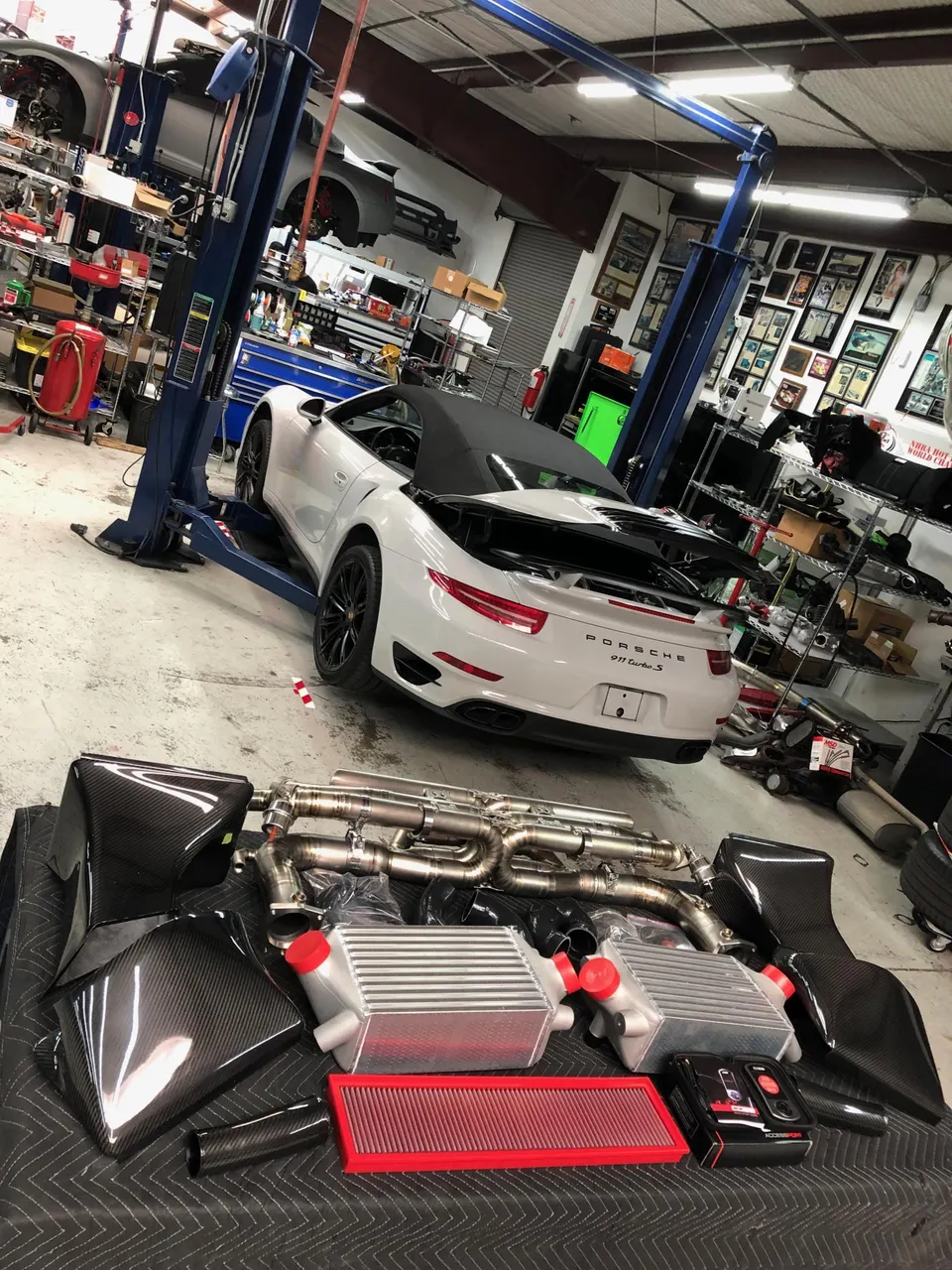 White Porsche 911 Turbo S on a lift in a performance shop with new intercoolers, exhaust, and carbon fiber intake parts laid out.