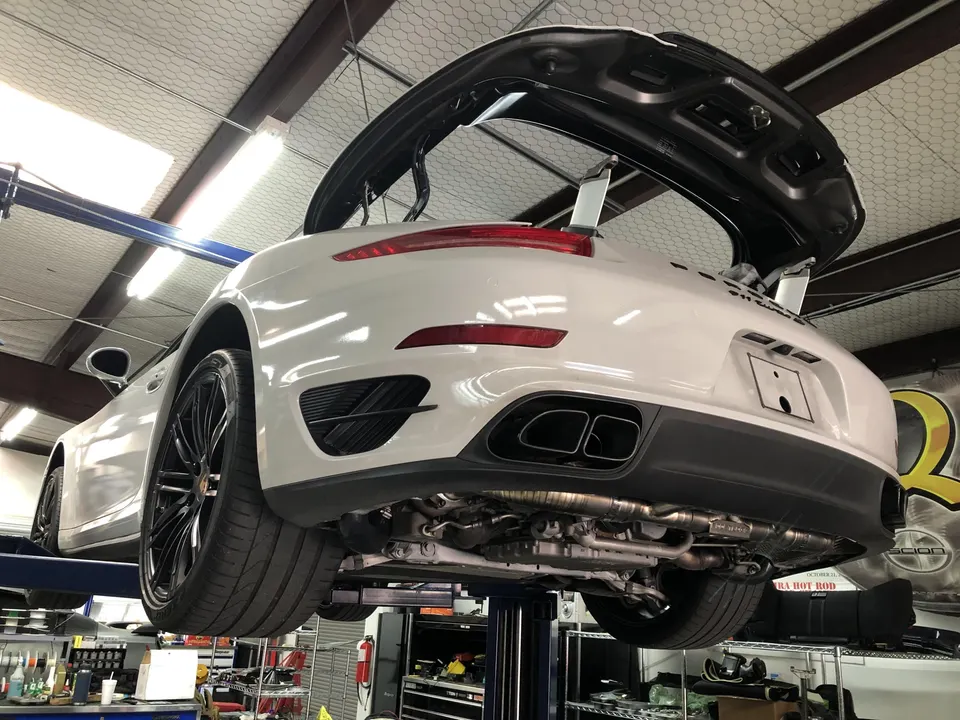 A white Porsche 911 Turbo S on a lift in a shop, with its rear engine cover open and the exhaust system visible.