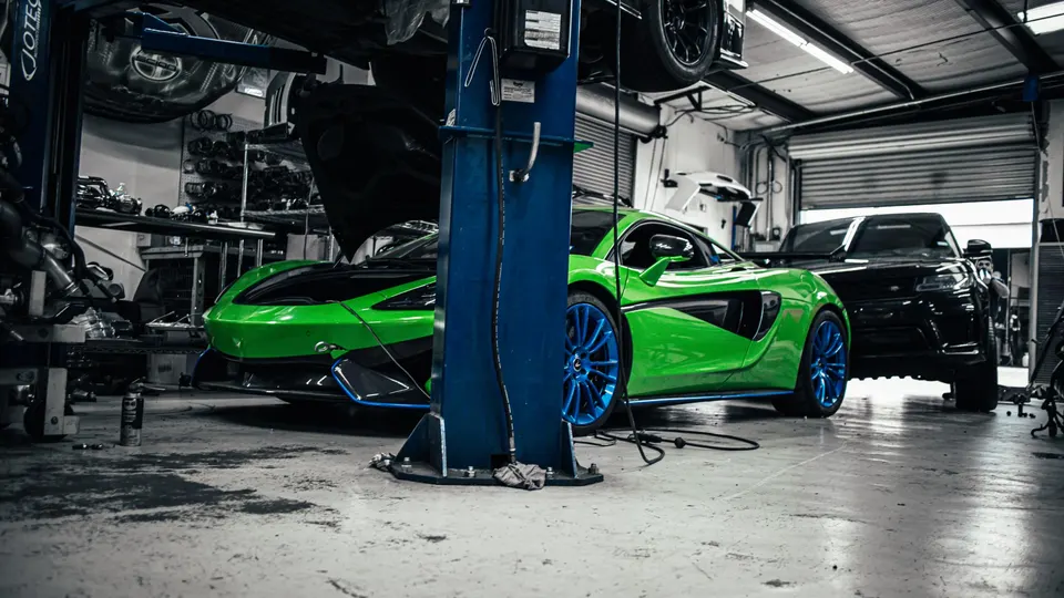 A vibrant green McLaren with striking blue wheels sits in a performance tuning shop, with another car on a lift.