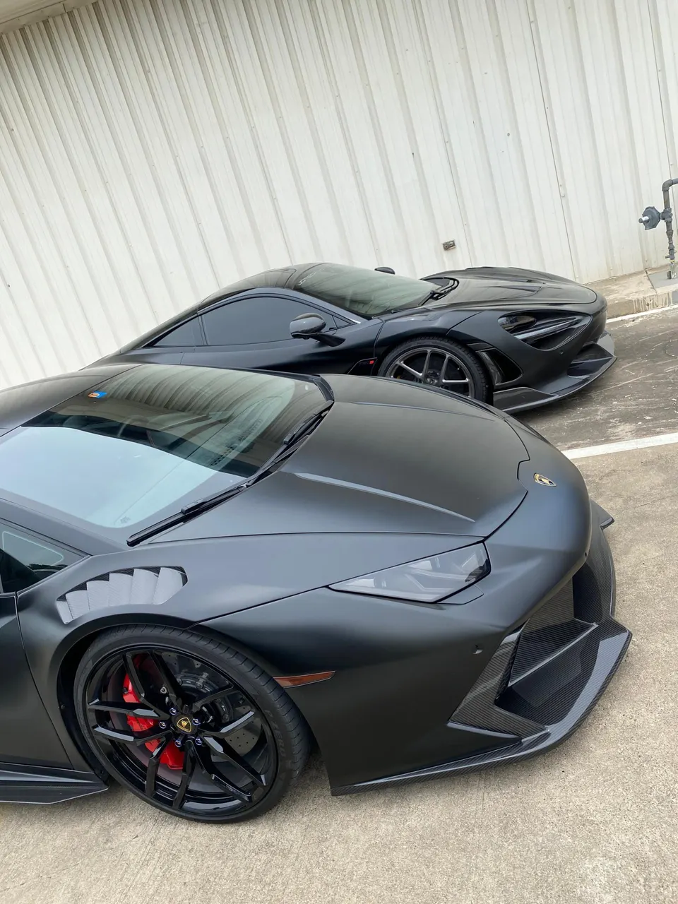 Two matte black supercars, a Lamborghini Huracan and a McLaren 720S, parked side-by-side, showcasing their aggressive front designs.