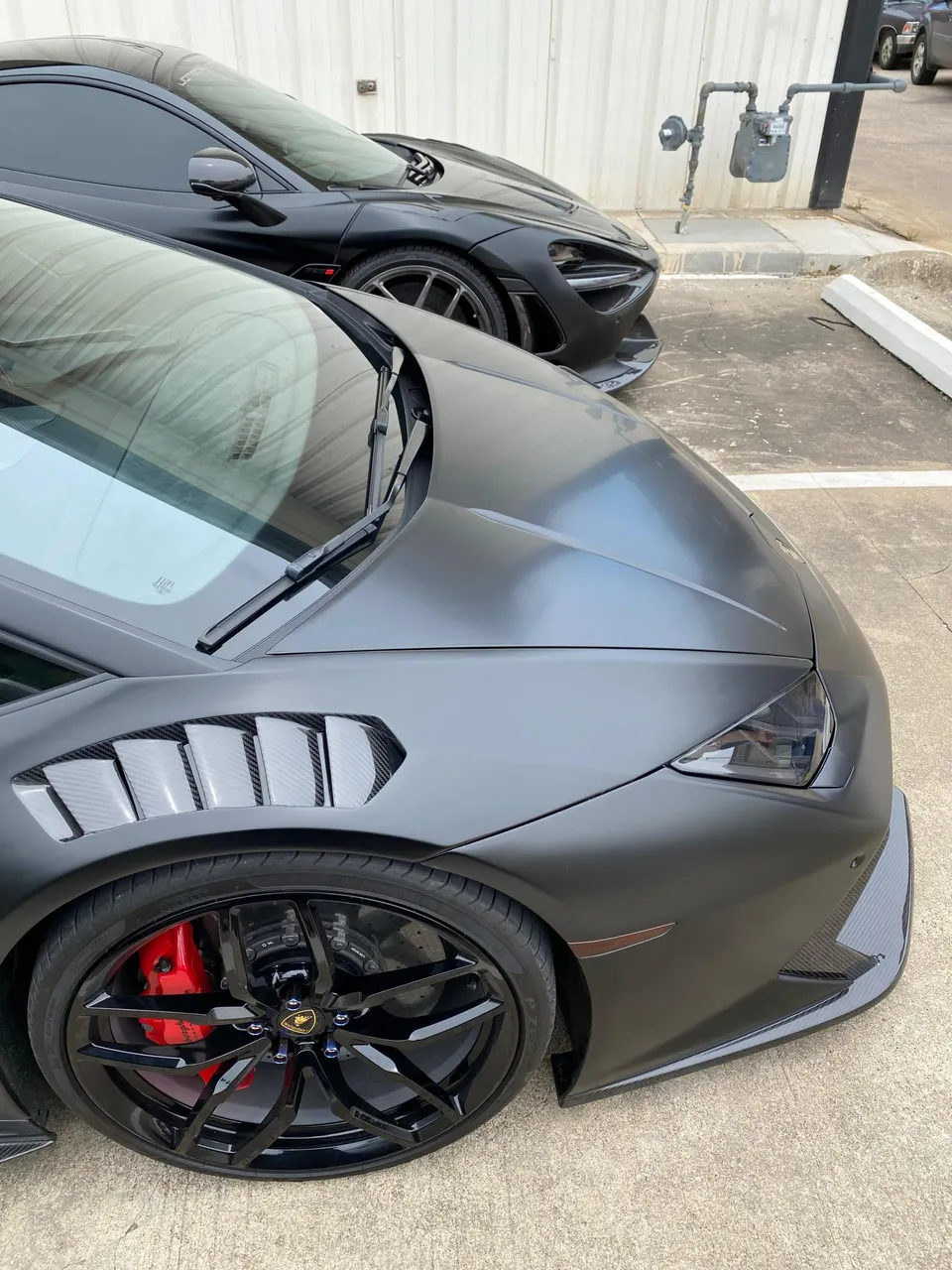 Matte black Lamborghini Huracan with carbon fiber fender vents and red brake calipers parked next to a McLaren 720S.