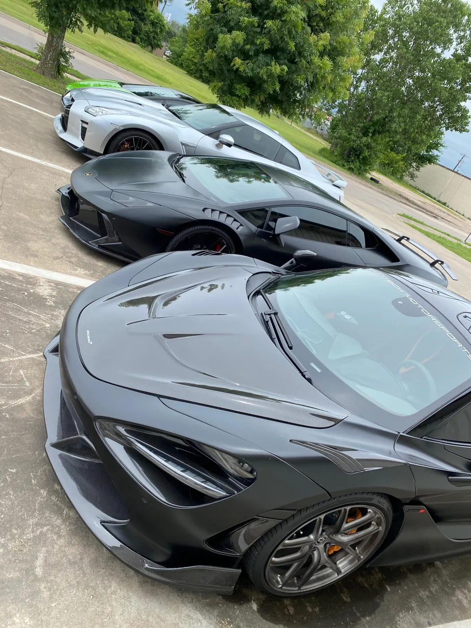 Three high-performance cars, a black McLaren, a matte black Lamborghini, and a silver Nissan GT-R, parked in a row.