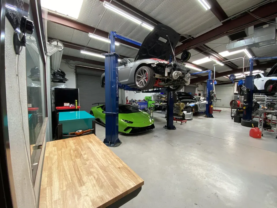 A performance tuning shop with a silver Toyota Supra on a lift showcasing a turbo setup, and a green Lamborghini Huracan on the floor.