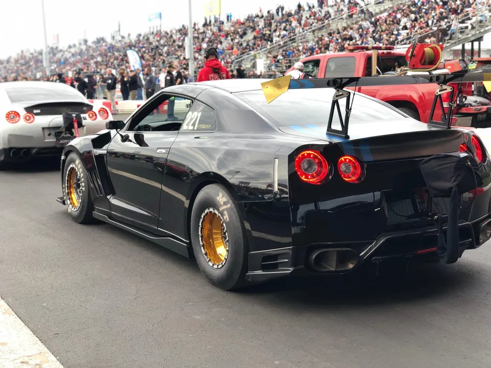 Black Nissan GT-R with widebody kit, large rear wing, and gold drag wheels in the staging lane at a drag strip.