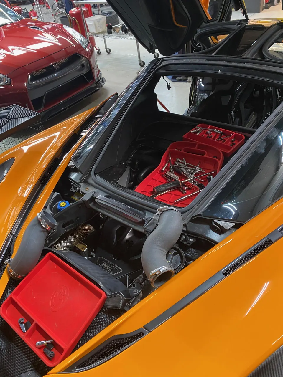 Orange McLaren 720S with its engine bay open and tools laid out in a performance tuning shop, with a red Nissan GT-R in the background.