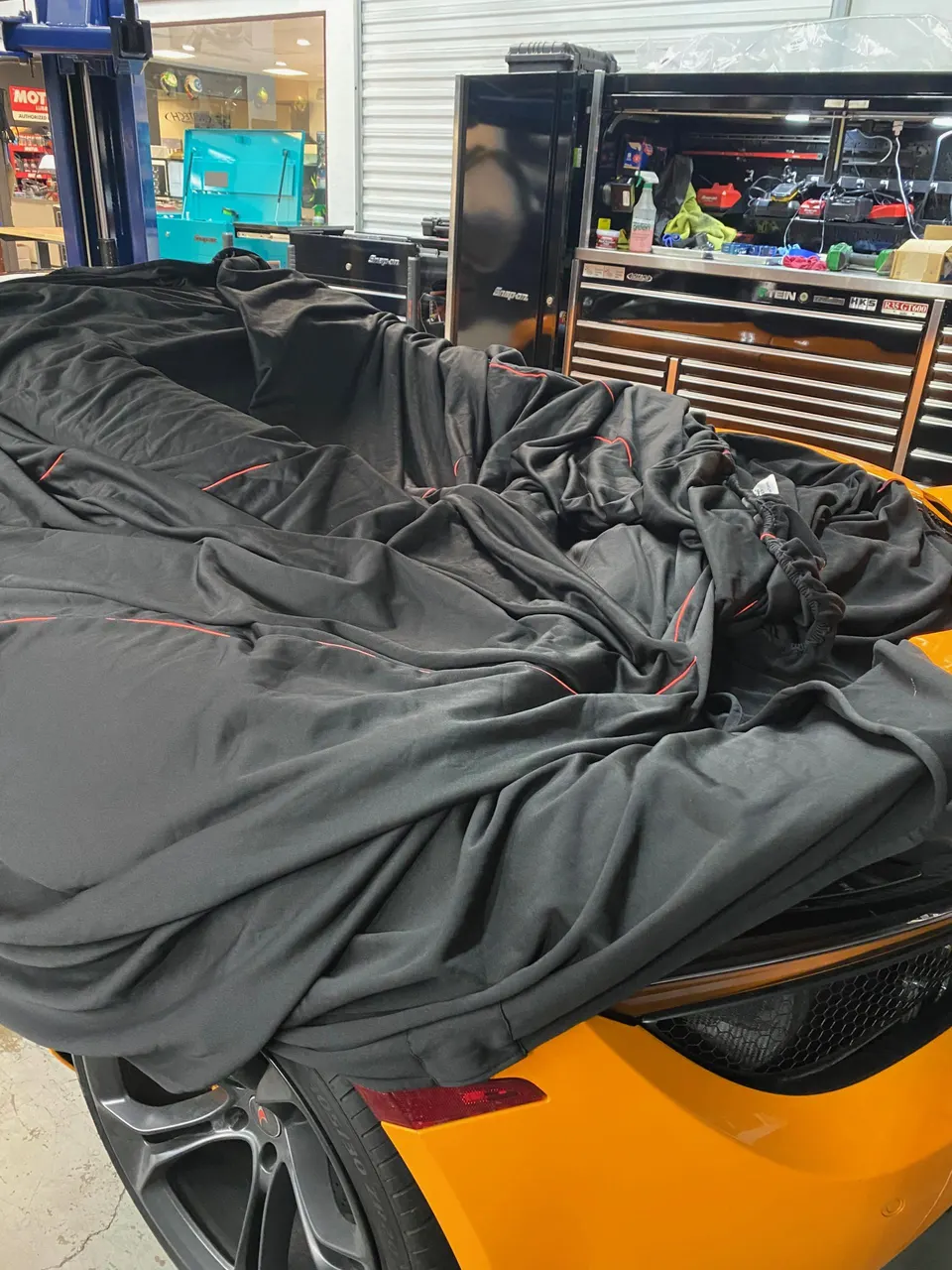 An orange McLaren sports car partially covered by a black car cover inside a Jotech Motorsports shop with toolboxes in the background.