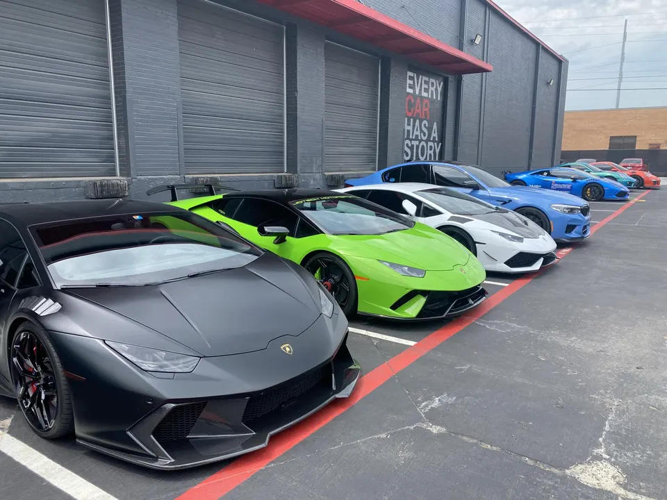 A lineup of high-performance supercars including a matte black Lamborghini Huracan, a green Lamborghini, and a blue BMW M5 parked outside a building.