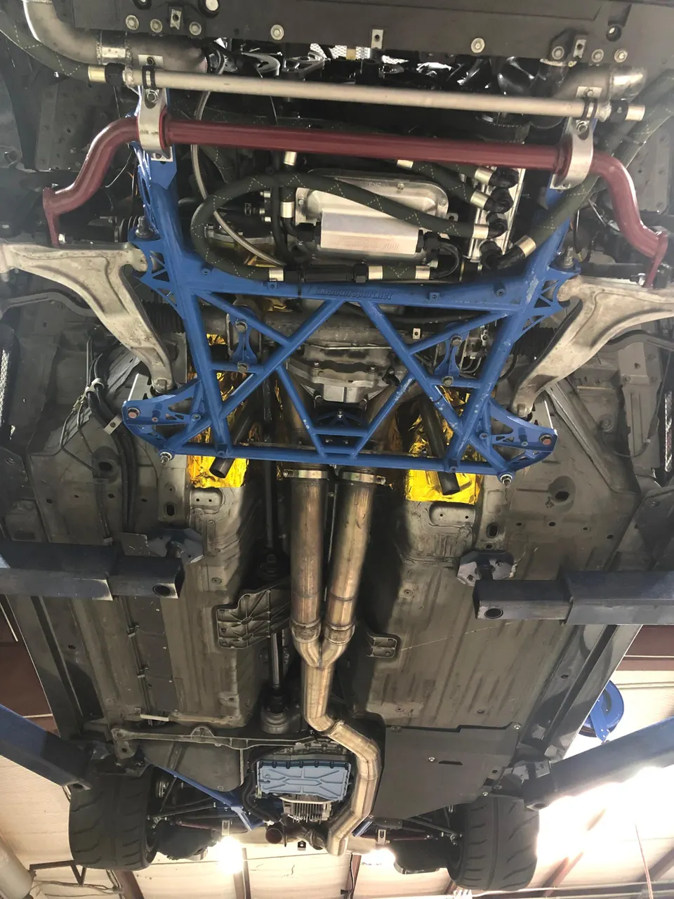 Underside view of a performance car on a lift, showcasing a blue chassis brace, aftermarket exhaust system, and fuel system components.
