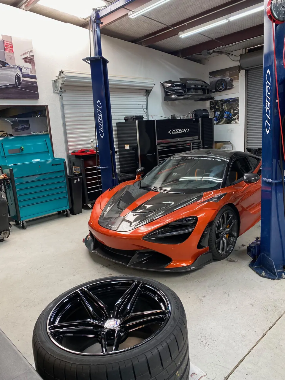 Orange McLaren 720S in a performance tuning shop with a new black multi-spoke wheel and tire in the foreground.